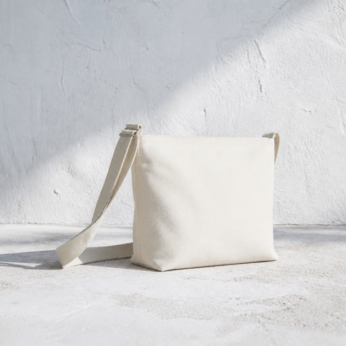 A cream canvas cross-body bag shot flat on a stone surface.