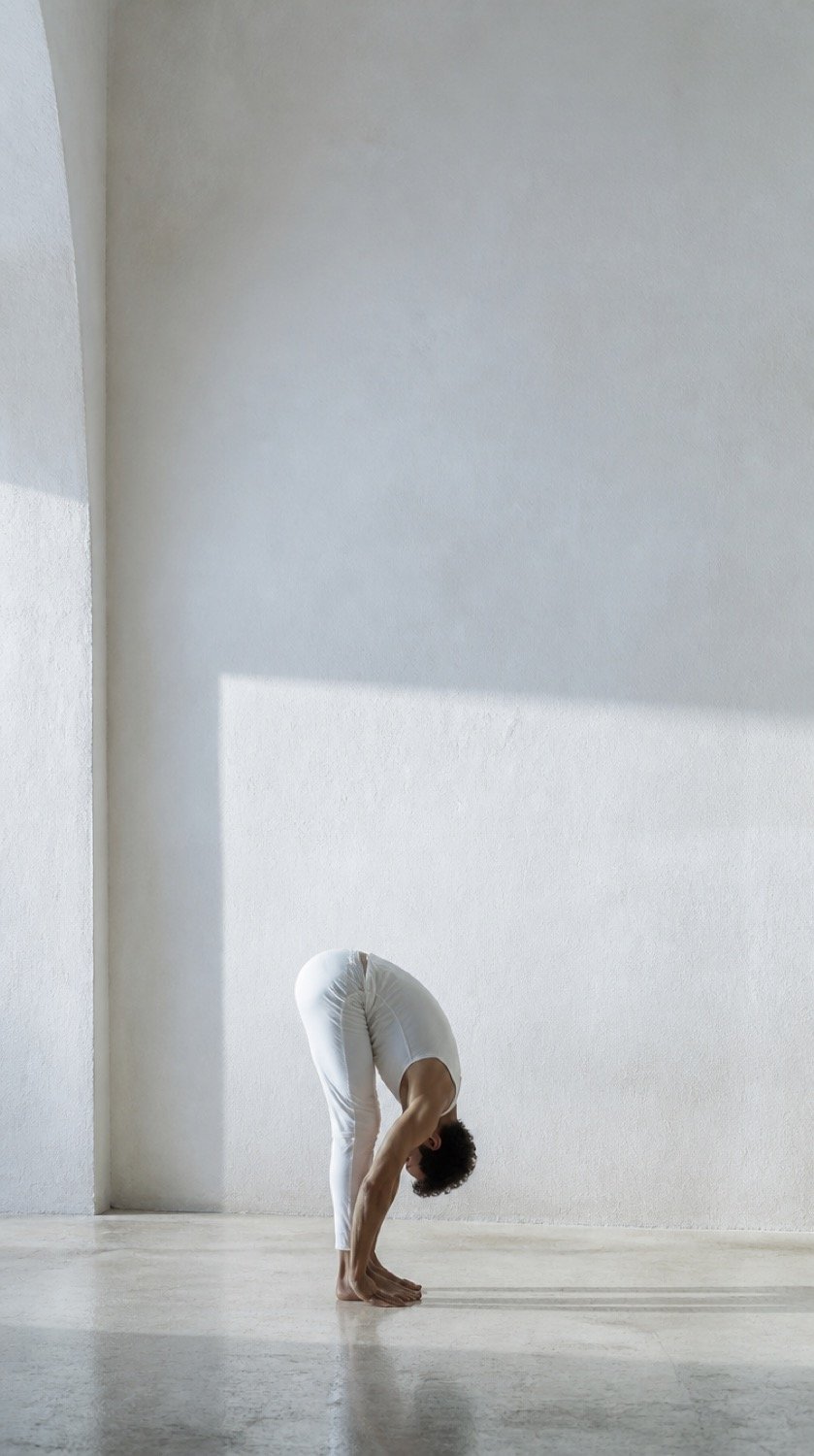 A standing forward fold performed in a wide, empty limewashed room.