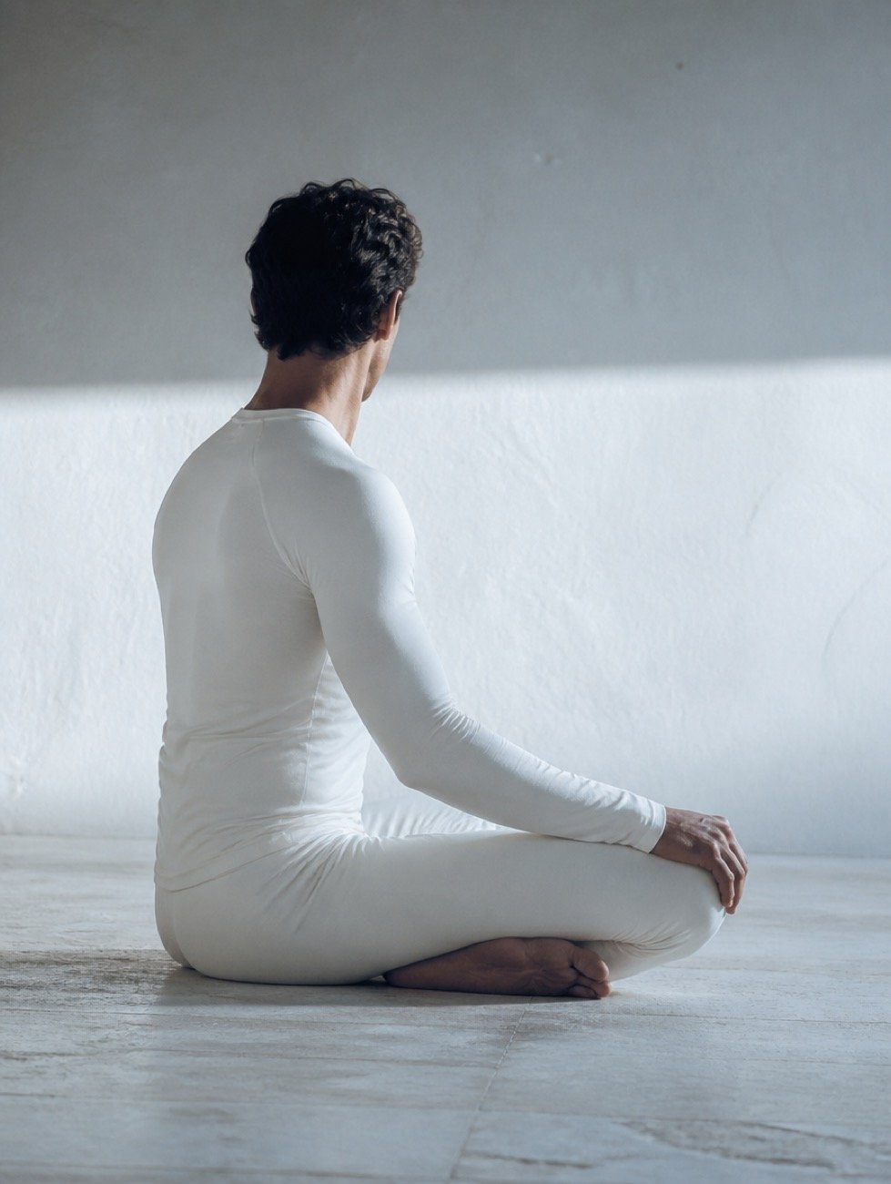 A seated meditation posture in a white long-sleeve Kinesis set, photographed from behind.