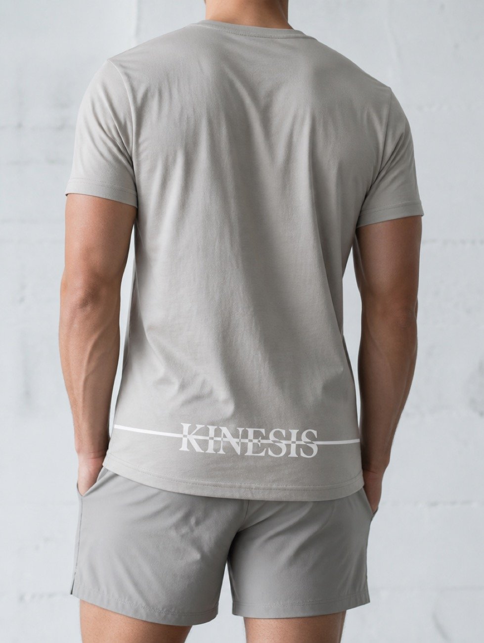 A men's grey Kinesis tee shown from behind, the wordmark and its white strike line running across the back above grey shorts.