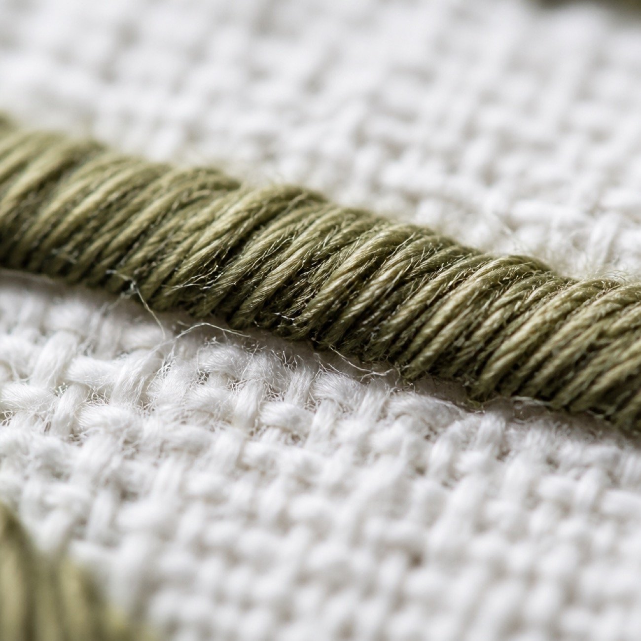 Macro photograph of olive thread stitching across a pale woven fabric.