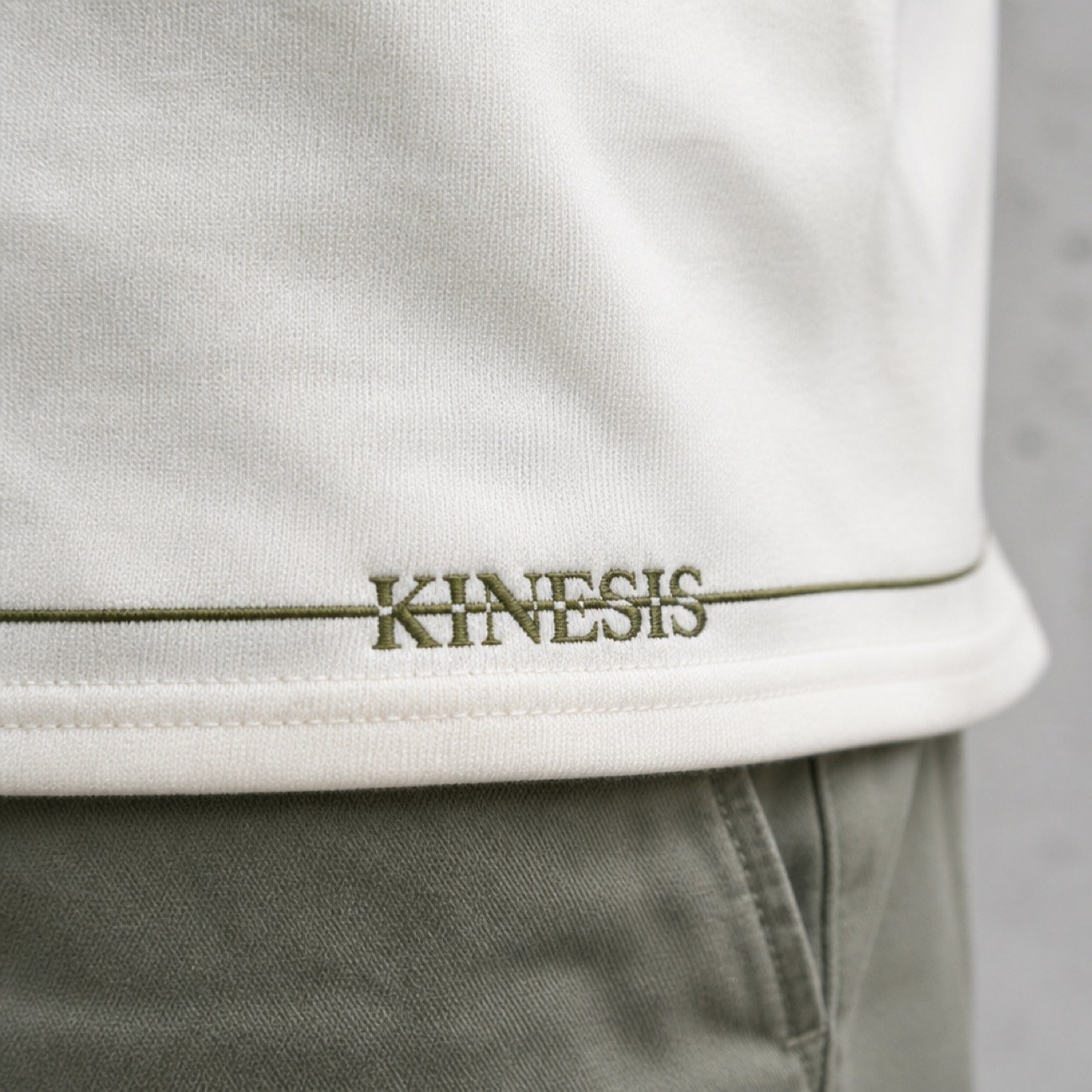 The Kinesis wordmark embroidered in olive at the hem of a cream tee, worn over olive trousers, the strike line running out across the fabric.