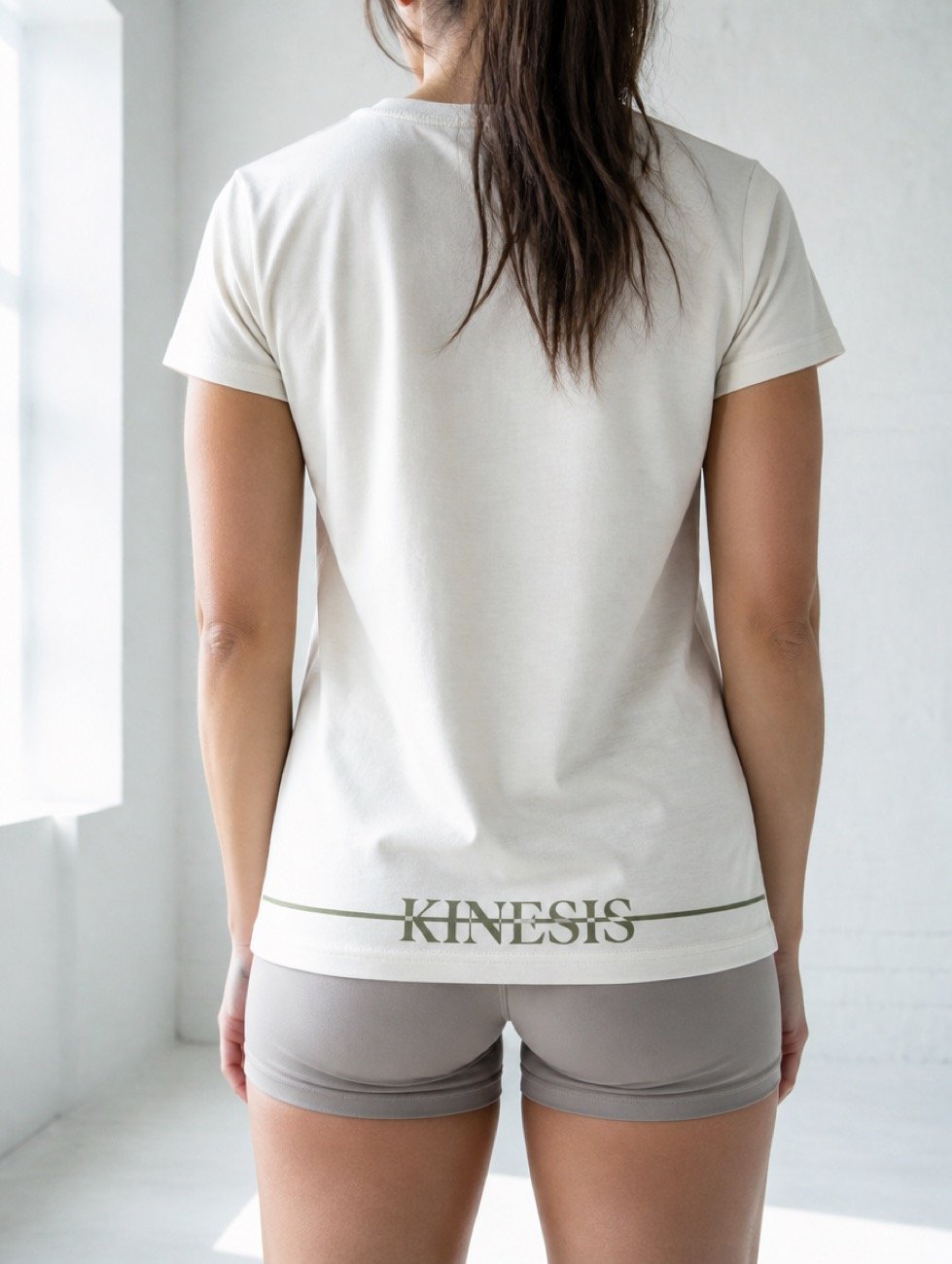 A women's cream Kinesis tee shown from behind, the wordmark and its olive strike line set across the hem above grey shorts.